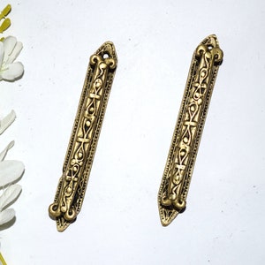 May include: Two antique brass-coloured cabinet pulls with ornate, symmetrical designs. Each pull is rectangular with a pointed end and a decorative handle. The pulls are set against a white background, suggesting a vintage or restoration theme.