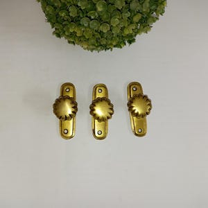 May include: Three vintage brass cabinet pulls with a flower-shaped knob. Each pull has a rectangular backplate with two screw holes. The pulls are a warm gold color and are arranged on a white surface.