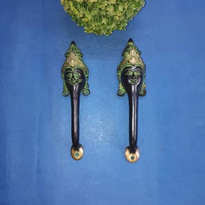 May include: Two ornate, dark-colored door handles with a face design. The handles have a brass-colored base and a detailed face with green accents. The handles are set against a blue background.