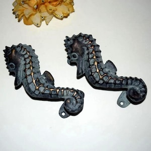 May include: Two seahorse-shaped decorative metal pieces with a distressed gray finish and gold accents. The seahorses have a detailed, textured design and are positioned side-by-side. A yellow flower is in the background.