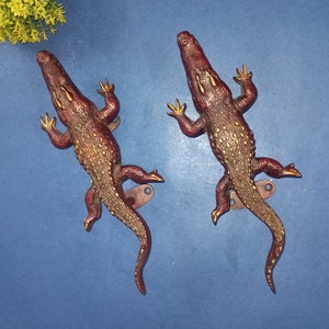 Könnte beinhalten: Zwei dekorative Krokodil-Wandhaken mit goldenen Akzenten. Die Haken sind aus Metall und haben eine rotbraune Oberfläche.