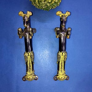 以下が含まれることがあります： キリンのようなデザインの装飾ドアハンドル2つ。ハンドルは黒色で、金色のアクセントと詳細な彫刻が施されています。青い表面に取り付けられており、背景には緑の植物があります。