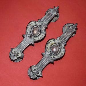 以下が含まれることがあります： 装飾的なディテールが施された、2つのアンティークブロンズ引き出しプル。各プルは、中央の円形デザインと、装飾的な要素とテクスチャードハンドルを備えています。プルは円形のバックプレートに取り付けられています。