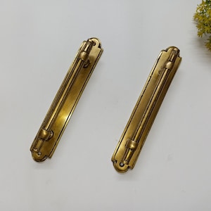 Op de afbeelding: Twee messingkleurige metalen kastdeurgrepen met een eenvoudig, rechthoekig ontwerp. De grepen hebben een licht gebogen vorm en een gladde afwerking.