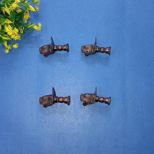 Pode incluir: Quatro puxadores de armário de cor bronze com um design de cabeça de cão. Os puxadores estão dispostos sobre uma superfície azul, com flores amarelas no canto superior esquerdo. Cada puxador tem uma cabeça de cão detalhada e uma base cilíndrica.