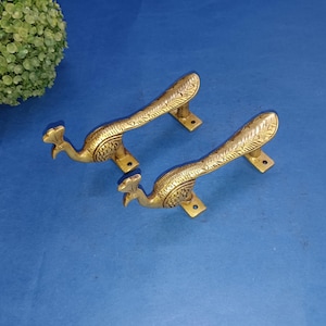 Puede incluir: Dos tiradores de puerta de pavo real de color dorado con detalles intrincados. Los tiradores tienen forma de pavo real con las colas extendidas.