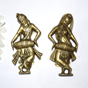 Op de afbeelding: Twee messingkleurige sculpturen van figuren die op trommels spelen. De figuren staan in een danshouding en dragen traditionele kleding. De sculpturen zijn op een wit oppervlak gemonteerd. De figuren zijn ongeveer 15 cm hoog.