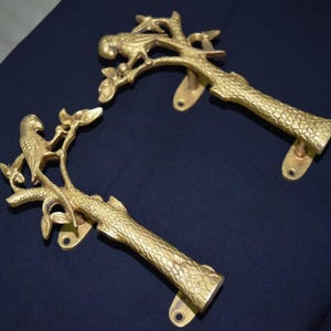 Op de afbeelding: Twee goudkleurige deurklinken. Elke klink toont een vogel die op een tak met bladeren zit. De klinken hebben een getextureerd, schorsachtig uiterlijk en zijn gemonteerd op een rechthoekige basis met schroefgaten.