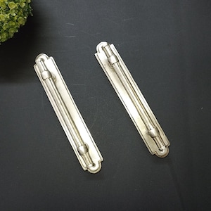 Op de afbeelding: Twee zilverkleurige kastgrepen met een rechthoekige achterplaat en een cilindrische handgreep. De grepen hebben een klassiek ontwerp met decoratieve einddetails. De hardware is waarschijnlijk voor meubels of kasten.