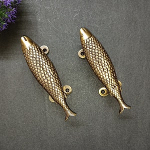 May include: A pair of bronze-coloured fish-shaped cabinet pulls. Each pull has a detailed scale pattern and a fin-shaped handle. The pulls are mounted on small, circular backplates with screw holes, designed for easy installation on furniture or doors.