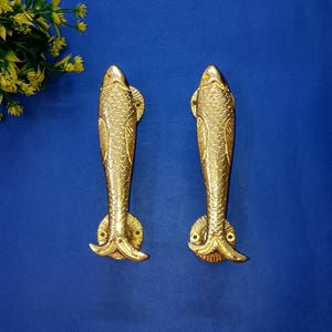 May include: Two gold-coloured, fish-shaped door handles. The handles have detailed scales and fins, set against a blue background. The handles are made of metal, likely for a door or cabinet.