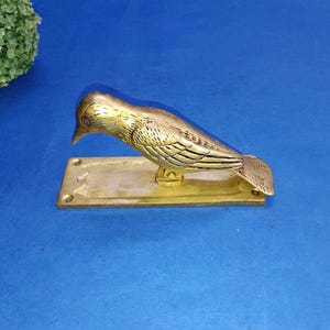 Puede incluir: Un llamador de puerta de latón dorado con forma de pájaro. El pájaro tiene detalles de plumas grabadas y está montado sobre una base rectangular. El llamador es de color dorado y se encuentra sobre un fondo azul.