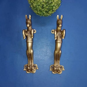 May include: Two brass door handles in the shape of a donkey. The handles are detailed with intricate carvings and have a vintage appearance. The donkey figures are standing upright, with their ears perked up. The handles are mounted on a blue surface.