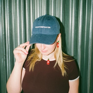 May include: A dark blue baseball cap with the text OUTFIT REPEATER embroidered in white on the front. The cap features a structured crown and a curved brim, constructed from a solid fabric with visible stitching details across the panels.