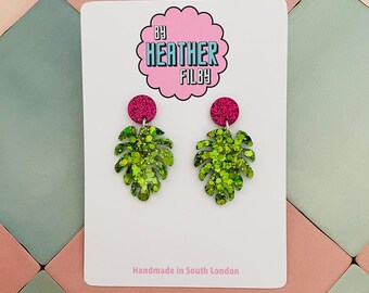 May include: A pair of leaf-shaped earrings with green glitter and pink accents. The earrings are attached to a pink circular stud. The earrings are on a white card with the text "By Heather Filby" and "Handmade in South London".