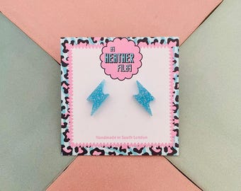 May include: A pair of light blue glitter lightning bolt earrings on a white card with a pink and black leopard print border. The card has the text "Heather Filby" and "Handmade in South London". The background is pink and green.