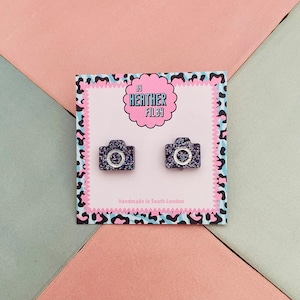 Puede incluir: Pendientes con forma de cámara con acabado azul oscuro brillante. Presentados en una tarjeta rosa con borde de estampado de leopardo y el texto "By Heather Filby" y "Handmade in South London".