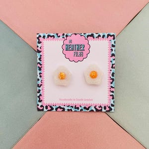 May include: Novelty fried egg earrings with white and glittery yellow centres, presented on a card. The card has a pink and black leopard print border, and the text "By Heather Filby" and "Handmade in South London".