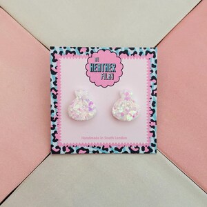 May include: A pair of white, glittery, money bag-shaped stud earrings displayed on a square card with a pink centre and a blue and black leopard print border. The card features the text By Heather Filby and Handmade in South London.