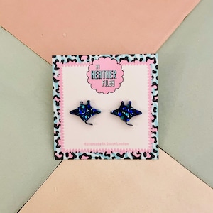 May include: A pair of dark blue, glittery stingray stud earrings. The earrings are on a card with a pink and black leopard print border. The card has the text "Heather Filby" in a pink bubble.
