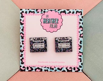 May include: A pair of black glittery cassette tape stud earrings displayed on a square card with a pink center and a black and blue leopard print border. The card features the text By Heather Filby and Handmade in South London.