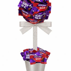 May include: A topiary-style arrangement featuring Cadbury Dairy Milk Biscoff chocolate bars in a spherical cluster atop a central stem, accented with a silver ribbon bow, and a base filled with more chocolate bars in a silver metal bucket.