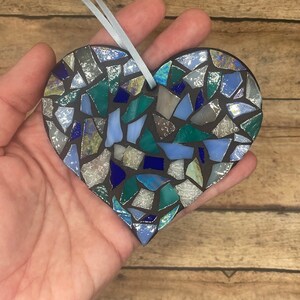 May include: A blue, green, and silver mosaic heart ornament. The heart is made of small, irregular pieces of glass and is attached to a light blue ribbon.