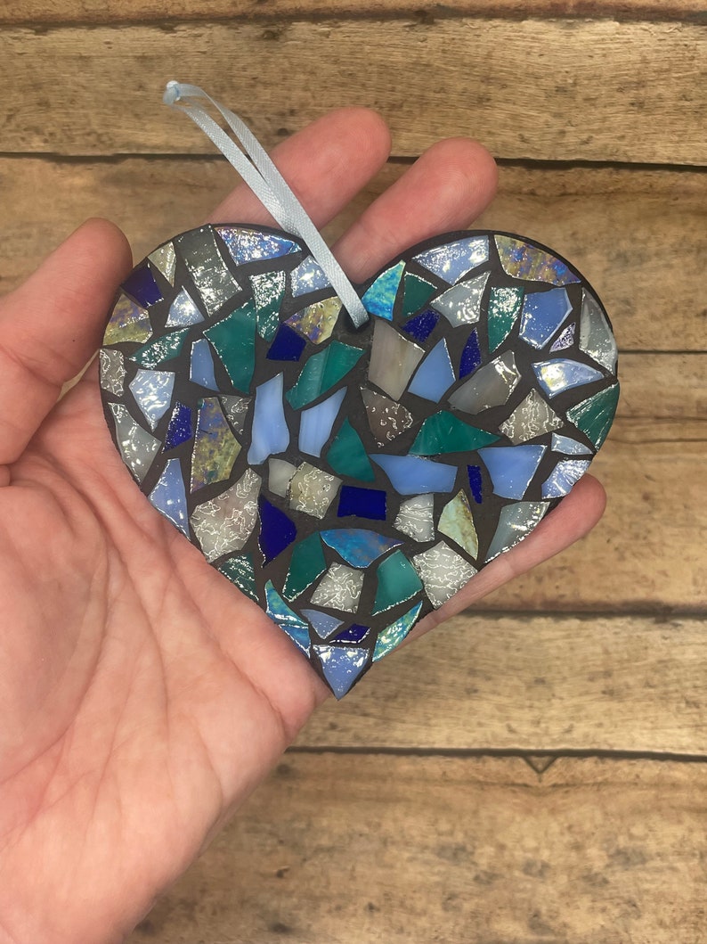 May include: A blue, green, and silver mosaic heart ornament. The heart is made of small, irregular pieces of glass and is attached to a light blue ribbon.