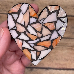 May include: A heart-shaped mosaic made of small, irregular pieces of glass in shades of white, orange, and pink. The mosaic is set in black grout.