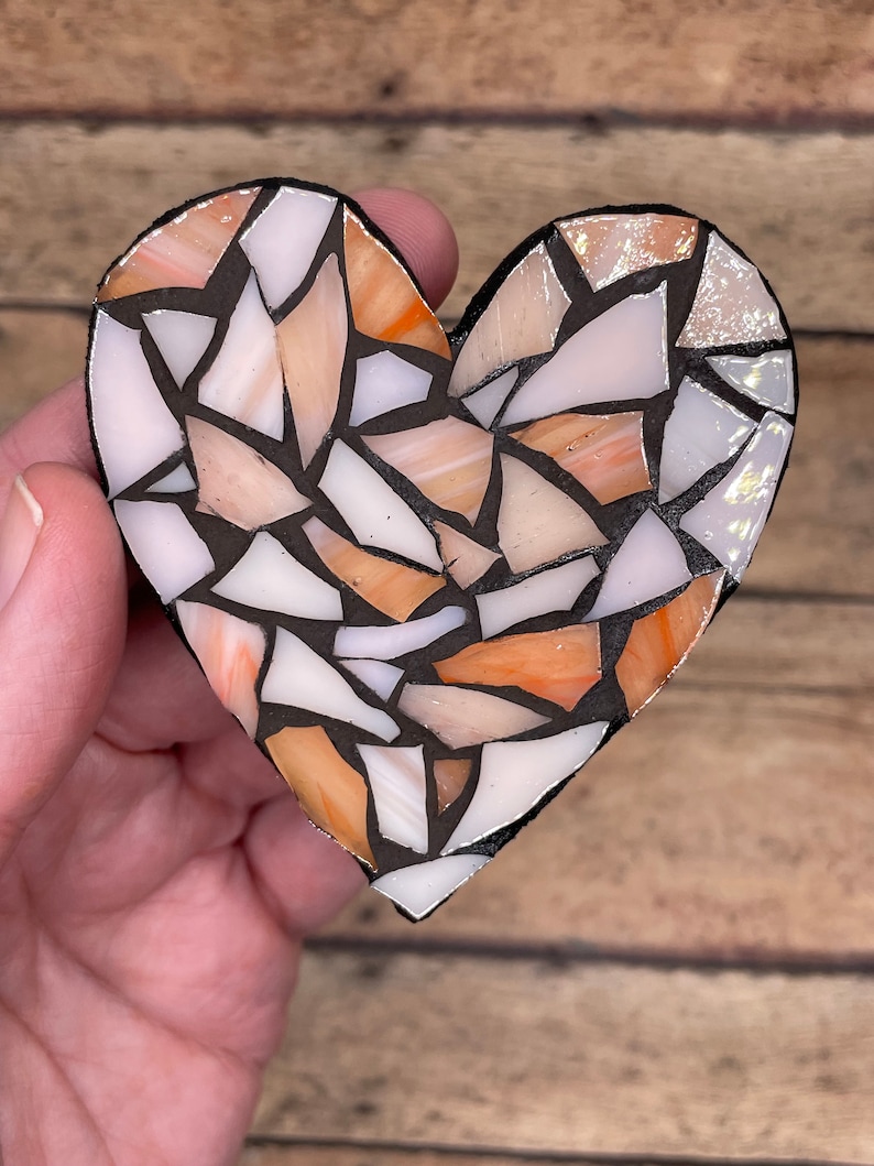 May include: A heart-shaped mosaic made of small, irregular pieces of glass in shades of white, orange, and pink. The mosaic is set in black grout.