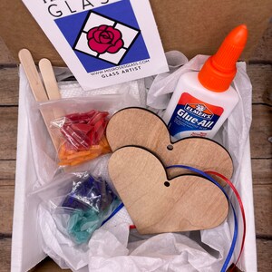 May include: A craft kit for making stained glass hearts. The kit includes two wooden heart shapes, two pieces of ribbon, a small bottle of glue, two wooden sticks, and two bags of glass pieces. The glass pieces are red, blue, and green.