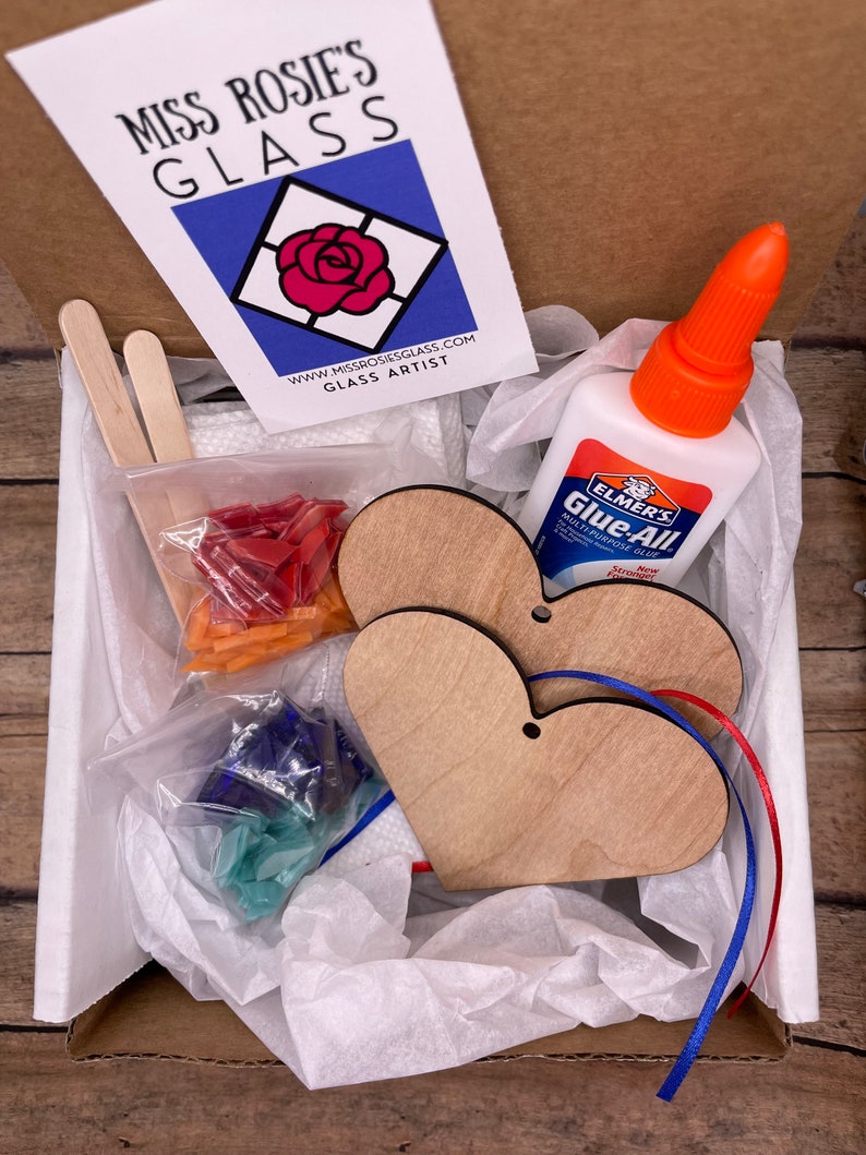 May include: A craft kit for making stained glass hearts. The kit includes two wooden heart shapes, two pieces of ribbon, a small bottle of glue, two wooden sticks, and two bags of glass pieces. The glass pieces are red, blue, and green.