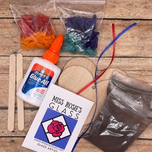 May include: A craft kit for making stained glass art. The kit includes two bags of colorful glass pieces, two wooden circles, a bottle of glue, two wooden sticks, a bag of brown powder, and a card with the text "Miss Rosie's Glass" and "www.missrosiesglass.com Glass Artist".