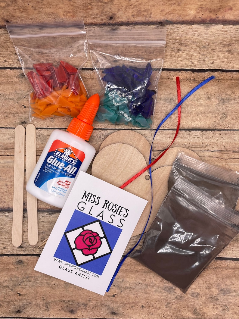 May include: A craft kit for making stained glass art. The kit includes two bags of colorful glass pieces, two wooden circles, a bottle of glue, two wooden sticks, a bag of brown powder, and a card with the text "Miss Rosie's Glass" and "www.missrosiesglass.com Glass Artist".
