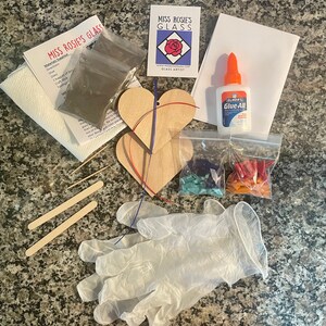 May include: A glass art kit with two wooden heart-shaped ornaments, glass pieces in blue, green, and orange, glue, and a pair of gloves. The kit also includes instructions and a small bag of glass glitter.