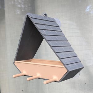 以下が含まれることがあります： 屋根のようなデザインのグレーとベージュのバードフィーダー。フィーダーは、テクスチャ加工された外観のグレーの傾斜した屋根とベージュの給餌トレイを備えています。3つの小さな止まり木がトレイの前面から伸びています。フィーダーは吸盤で窓に取り付けられています。