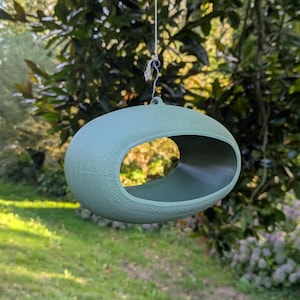 Puede incluir: Un comedero para pájaros ovalado de color verde azulado apagado, colgado de una rama. El comedero tiene una gran abertura y está suspendido por un fino alambre. El fondo presenta follaje verde y un césped.