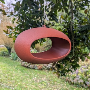 Puede incluir: Un comedero para pájaros de forma ovalada, de color terracota, con una gran abertura, suspendido de la rama de un árbol. El comedero tiene una superficie texturizada y se encuentra sobre un fondo borroso de follaje verde y césped.