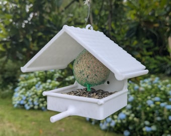 May include: A white bird feeder with a pitched roof, hanging from a string. The feeder holds a suet ball and seeds. The background features green foliage and blue flowers.