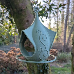 Può includere: Un mangiatoia per uccelli verde salvia con un design a spirale decorativo, appeso a un ramo d'albero. L'alimentatore ha un tetto a punta e un posatoio circolare. Lo sfondo mostra una scena di foresta sfocata.