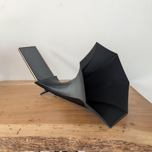 Op de afbeelding: Een matzwart, sculpturaal object met een uitlopende, hoornachtige opening en een rechthoekige basis. De vorm heeft gladde, gebogen oppervlakken die overgaan van een smalle hals naar een brede, veelzijdige, waaiervormige opening.
