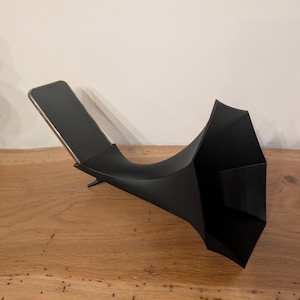 Puede incluir: Un soporte de teléfono de metal negro con un diseño curvo único. El soporte sostiene un teléfono inteligente, inclinado hacia arriba. El soporte está sobre una superficie de madera, con una pared blanca de fondo.