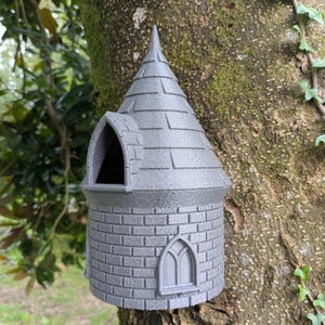 May include: A grey, castle-shaped birdhouse with a conical roof and arched window. The birdhouse is mounted on a tree trunk, blending with the natural surroundings. The birdhouse is made of a brick-like material.