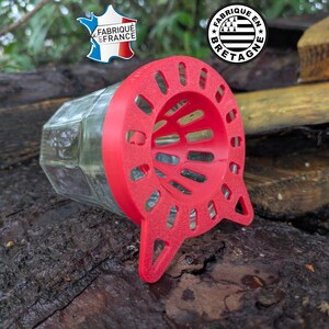 Peut inclure: Un bocal en verre transparent à corps hexagonal avec un couvercle en plastique rouge vif doté d'une large ouverture en forme d'entonnoir avec des fentes intégrées. Le couvercle présente deux petites languettes à la base. Deux logos sont visibles.