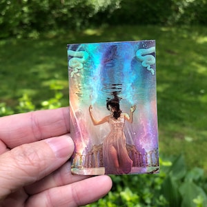 May include: A rectangular art print featuring a woman in a pink dress submerged in water, with a celestial background. The image has a holographic effect, with blue and purple hues. Two serpent-like figures are at the top.