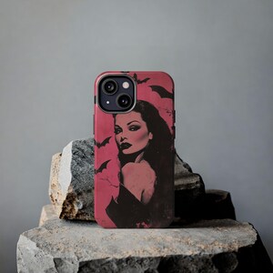 May include: A phone case featuring a vintage-style illustration of a woman with dark hair and red lipstick, set against a pink background with bat silhouettes. The case is black and pink.