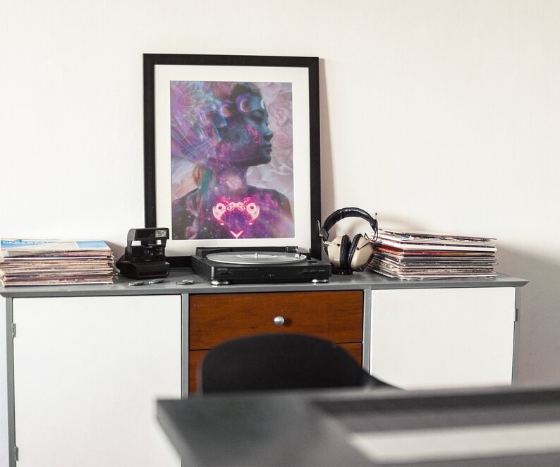 Op de afbeelding: Een ingelijste print van een vrouwen gezicht met een galaxie achtergrond. De print staat op een plank met een platenspeler, koptelefoon en een stapel vinyl platen.