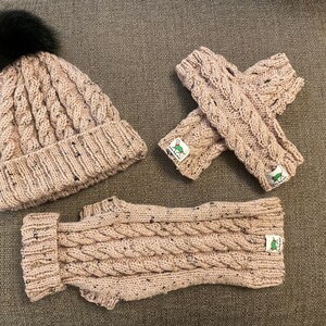 May include: A set of knitted winter accessories in a light brown colour. The set includes a beanie with a black pom pom, fingerless gloves, and a neck warmer. All items feature a cable knit pattern.