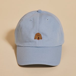 Può includere: Un berretto da baseball blu chiaro con un viso di cane marrone ricamato sul davanti.
