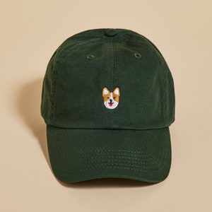 K&ouml;nnte beinhalten: Eine dunkelgr&uuml;ne Baseballkappe mit einem kleinen gestickten Bild eines Corgi-Hundes auf der Vorderseite.